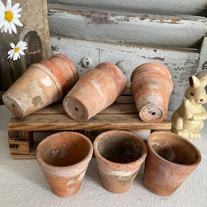 May include: A collection of terracotta flower pots, some overturned, on a small wooden pallet. The pots vary in size and show signs of weathering. A small, cream-colored bunny figurine sits nearby, adding a touch of whimsy to the rustic scene.