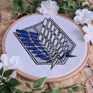 Puede incluir: Un aro de bordado blanco con un diseño de punto de cruz azul y gris del símbolo de Ataque a los Titanes. El símbolo es un emblema alado con forma de escudo con un contorno negro.