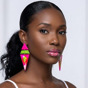 May include: Bright pink and green geometric earrings. The earrings have a diamond shape with a green horizontal stripe and abstract designs. The woman in the image is wearing a white tank top.