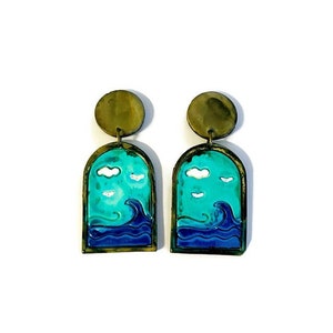 May include: A pair of artisan earrings featuring a sea-themed design. Each earring has a brass-colored top and a rectangular pendant with a turquoise sky, white clouds, and a dark blue ocean wave. The earrings are set against a white background.