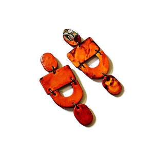 May include: A pair of orange earrings with a geometric design. Each earring features a rectangular top, a curved middle section, and a small oval at the bottom. The earrings are connected with black metal accents and have a silver-colored stud.