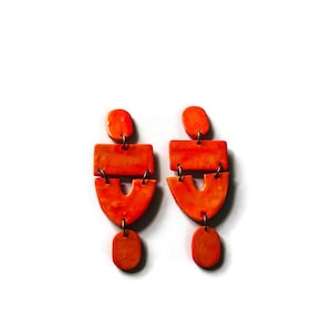 May include: A pair of bright orange dangle earrings featuring a three-part construction. Each earring consists of a small oval top, a rectangular middle section, and a U-shaped bottom piece, all connected by small metal jump rings in a vertical line.
