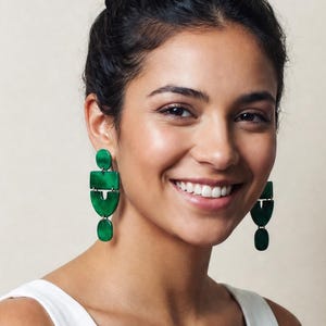 May include: A pair of emerald green drop earrings featuring a stacked geometric design with a rectangular top piece, a semi-circular middle section, and a small oval base, worn with a sleeveless white top featuring a deep V-neckline.