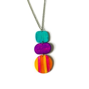 May include: A vibrant pendant necklace with a silver chain. The pendant comprises three stacked shapes: a turquoise square, a purple oval, and a striped orange and pink circle. The necklace is set against a white background.