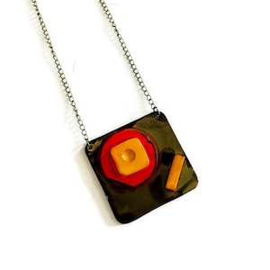 May include: A unique necklace featuring a square pendant with a dark metallic finish. The pendant has a red and yellow circular design with a small square and a rectangular accent. The necklace hangs from a delicate chain.