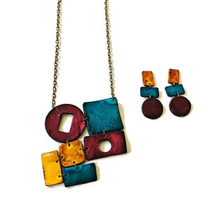 Op de afbeelding: Een geometrische ketting- en oorstekerset met turquoise, bordeauxrode en oranje accenten. De ketting heeft een cirkel, vierkanten en rechthoeken. De oorbellen zijn druppelvormig met een vierkant en een cirkel.