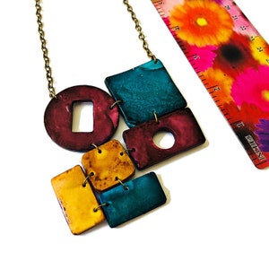 Op de afbeelding: Een kleurrijke geometrische ketting met turquoise, bordeauxrode en gele accenten. De ketting is gemaakt van polymer clay en heeft verschillende vormen, waaronder cirkels, vierkanten en rechthoeken.
