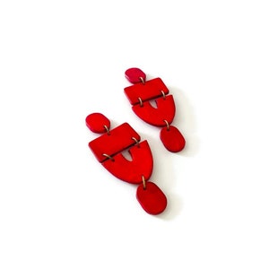 May include: A pair of bright red, geometric earrings. Each earring features a small, round stud, a rectangular piece, a curved shape with a cutout, and an oval drop. The pieces are connected with small gold-coloured metal rings.