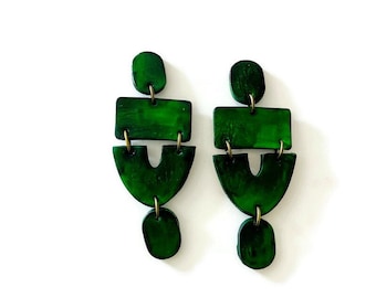 May include: A pair of dark green drop earrings featuring a vertical arrangement of three distinct shapes: a small oval top, a rectangular middle piece, and a U-shaped bottom section, all connected by small gold-toned metal jump rings.