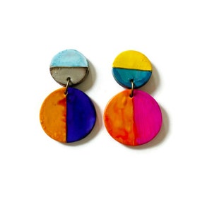 May include: A pair of colorful dangle earrings with two round discs per earring. The top disc is a light blue, teal, and gray color combination. The bottom disc is a bright orange and purple color combination on one earring and a bright orange and pink color combination on the other earring.