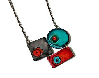 Op de afbeelding: Een ketting met een bronzen ketting en drie geometrische hangers. De hangers zijn geschilderd in tinten grijs, turquoise en rood met oranje accenten.