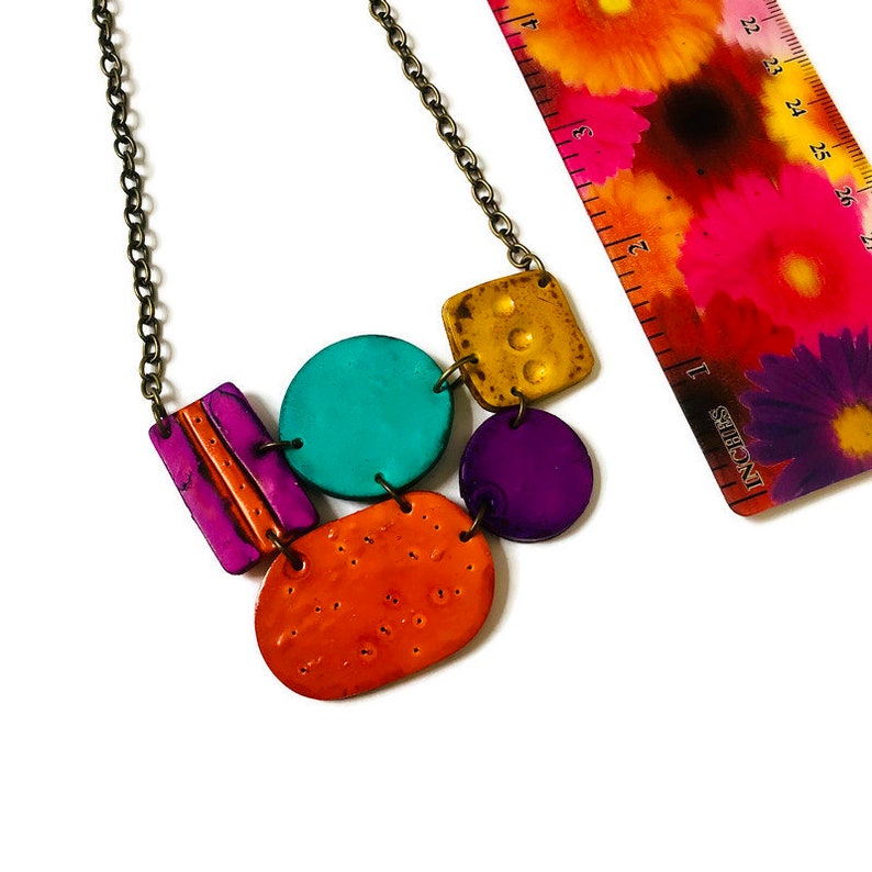 May include: A colorful necklace with geometric shapes made of polymer clay. The necklace features a teal circle, a purple circle, a yellow square, an orange oval, and a pink rectangle. The necklace is made with a bronze chain.