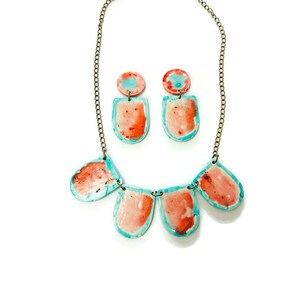May include: A necklace and earring set with a distinctive design. The jewellery features orange, blue, and white pendant shapes, connected by a chain. The earrings are small circles with a rectangular shape below. The set is handmade.