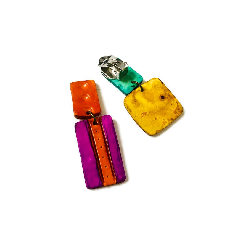 May include: A pair of colorful dangle earrings with a clip-on closure. The earrings are made of layered squares of orange, purple, yellow, and turquoise.