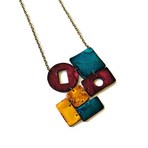 Op de afbeelding: Een geometrische ketting met een gouden ketting en kleurrijke hars hangers. De hangers zijn in tinten teal, bordeauxrood en geel.