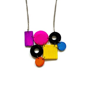 May include: A colorful geometric necklace with a chain, featuring a pink circle, a yellow square, a black circle with a hole in the center, a purple rectangle, a blue circle, and an orange circle.