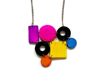 Op de afbeelding: Een ketting met een zilverkleurige ketting verbonden aan een cluster van geometrische vormen, waaronder een paarse rechthoek, een felroze cirkel, een zwarte ring, een geel vierkant met verticale lijnen, een kleine blauwe cirkel en een oranje cirkel.