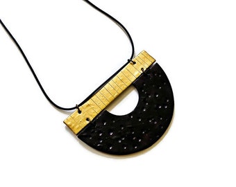 May include: A modern necklace with a black, semi-circular pendant featuring small, evenly spaced holes. The pendant is connected to a light-colored, rectangular section with vertical lines. The necklace hangs from a black cord.
