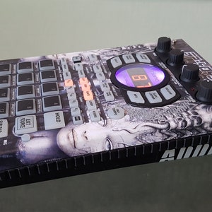 May include: A black and gray electronic drum machine with a graphic design of a face on the front. The machine has a digital display that reads "81". The text "055CHOT ULTRA 320" is printed on the top right corner of the machine.