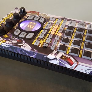Op de afbeelding: Een zwarte Roland SP-404SX sampler met een aangepast ontwerp met een witte en blauwe robot-illustratie. De sampler heeft een groot display, 16 pads en verschillende knoppen en draaiknoppen.