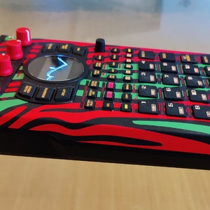 Peut inclure: Un échantillonneur de musique rouge, vert et noir avec le texte "055CHOT ULTRA 320" dans le coin supérieur droit. L'échantillonneur possède une variété de boutons et de boutons, y compris un grand bouton central.