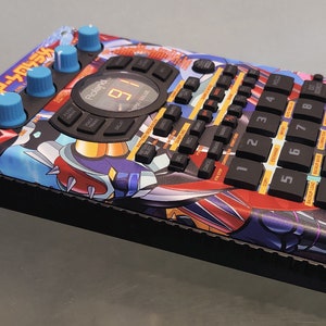 Peut inclure: Un échantillonneur Roland SP-404SX noir avec un design personnalisé représentant une illustration de style anime bleu, rouge et jaune. L'échantillonneur possède un écran numérique, 12 boutons et quatre boutons.