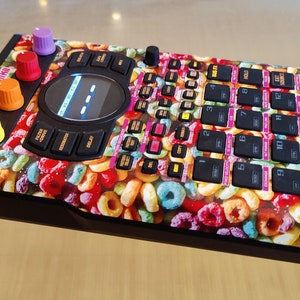 May include: A black and orange electronic music sampler with a colourful cereal design. The sampler has a large knob, a screen, and 16 buttons.