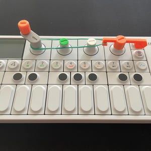 Peut inclure: Un synthétiseur blanc avec un clavier noir et blanc, des boutons noirs et des boutons orange et verts. Le synthétiseur est étiqueté "OP-1".