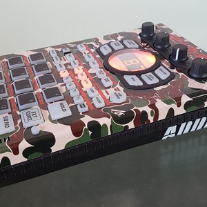 May include: A black and white camo-patterned Roland SP-404SX sampler with the text "ADDICT" on the side. The sampler has a red and black display and a red and black button layout.
