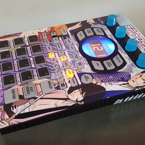 Puede incluir: Una caja de ritmos electrónica negra y azul con un diseño de anime colorido. La caja de ritmos tiene una pantalla que muestra "12" y tiene varios botones y perillas. El diseño presenta personajes de la serie de anime "Lupin III".