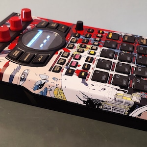 May include: A black and red music sampler with a colourful graphic design. The sampler has a large knob in the centre, a screen, and a grid of buttons. The text "055CHOT ULTRA 320" is visible in the top right corner.