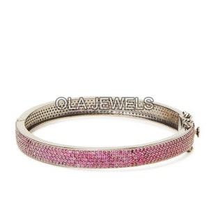 Op de afbeelding: Zilverkleurige armband met een rij kleine, levendige roze edelstenen. De armband heeft een scharnierende sluiting en de tekst "OLA JEWELS" is zichtbaar. Een sieraad.