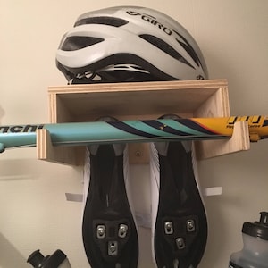 May include: A wooden bike storage rack with a white helmet, two pairs of cycling shoes, and two water bottles. The rack has a shelf for the helmet and hooks for the shoes and bottles.