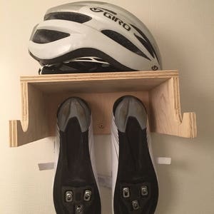 May include: A wooden wall-mounted bike storage rack with a white helmet and a pair of white cycling shoes with black cleats hanging on it. The rack has two hooks for hanging shoes and a shelf for storing a helmet.