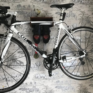 May include: A white Colnago road bike with black accents is mounted on a wall with a wooden shelf above it. The shelf has the word "Extreme" written on it. The bike has a black seat and black handlebars. The bike is on a white wall with a black and white animal print wallpaper.