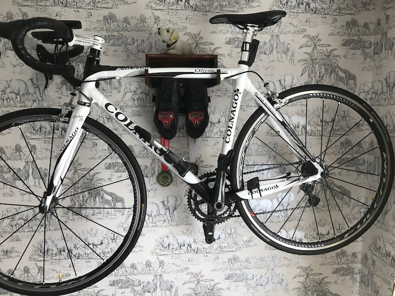 May include: A white Colnago road bike with black accents is mounted on a wall with a wooden shelf above it. The shelf has the word "Extreme" written on it. The bike has a black seat and black handlebars. The bike is on a white wall with a black and white animal print wallpaper.