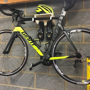 May include: A black and yellow Giant road bike is hanging on a wall with a helmet and cycling shoes. The bike has a black frame with yellow accents and black wheels with white tires. The helmet is yellow and the shoes are black.