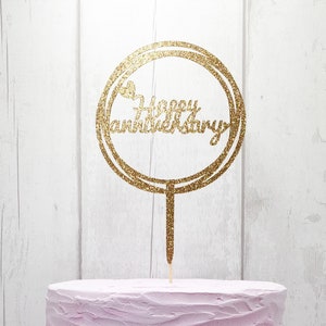 May include: Gold glitter cake topper with the words "Happy Anniversary" in a circle. The topper is on a pink frosted cake.