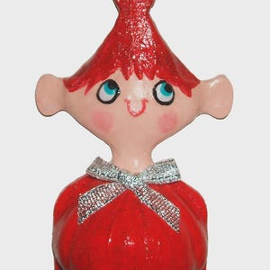 May include: A small figurine featuring a character with a pointed red hat, light pink skin, and large blue eyes. The character wears a red garment adorned with a silver glittery bow at the neckline. The figure has prominent ears and a small smile.