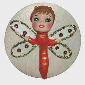May include: A whimsical, handcrafted dragonfly ornament. The ornament features a red body with gold stripes, a matching red hat, and blue-eyed face. The wings are light blue with gold glitter trim and red sequins, set against a white, glittery background.