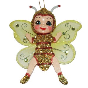 May include: A hanging ornament shaped like a bee character with a glittery gold and red striped body, a smiling face with rosy cheeks, and large, pale yellow translucent wings decorated with silver wire swirls and small clear rhinestones.