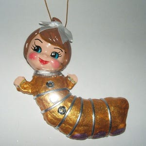 May include: A hanging ornament shaped like a caterpillar with a human-like face, featuring brown hair with a white bow, blue eyes, and rosy cheeks. The body is gold with silver ribbon accents, small sequins, and purple details at the base.