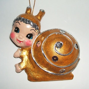 May include: A decorative snail ornament featuring a human-like face with rosy cheeks, large blue eyes, and long antennae. The snail has a metallic gold body and a spiral shell accented with silver glitter trim and scattered round silver sequins.