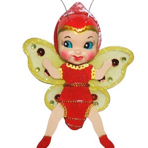 May include: A whimsical ornament featuring a smiling doll dressed as a butterfly. The doll has a red outfit, yellow glittery wings, and blue eyes. The ornament has two silver antennae with white pearl tips. A charming decorative piece.