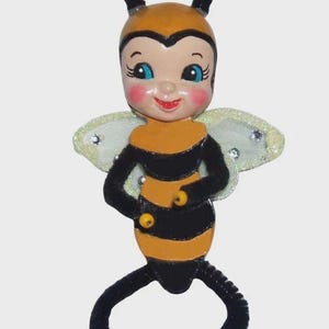 May include: A bee-shaped hanging ornament featuring a yellow and black striped body, a smiling face with blue eyes and rosy cheeks, two antennae with yellow tips, and glittery translucent wings. The limbs are made of black pipe cleaners.