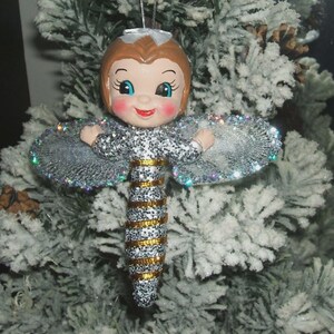 May include: A Christmas tree ornament shaped like a fairy with a doll-like face, brown hair, and rosy cheeks. The body is covered in silver glitter with horizontal gold stripes. It features wide, iridescent, glittery wings and a silver string.