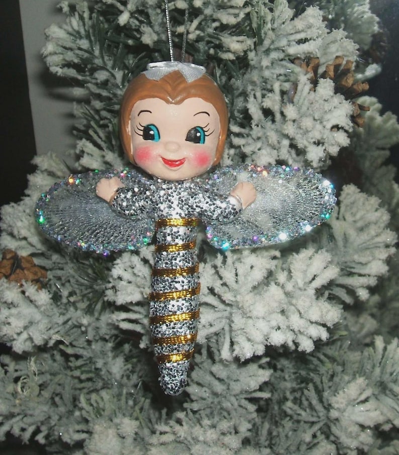 May include: A Christmas tree ornament shaped like a fairy with a doll-like face, brown hair, and rosy cheeks. The body is covered in silver glitter with horizontal gold stripes. It features wide, iridescent, glittery wings and a silver string.