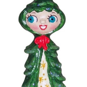May include: A whimsical, hand-painted figurine of a stylized Christmas tree person. The figure has a green, textured exterior with a red bow and a white dress adorned with gold stars. The face has blue eyes and a smiling expression.