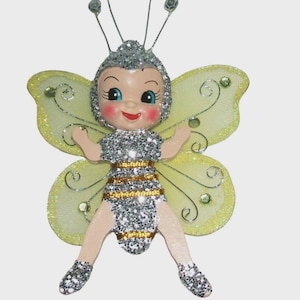 May include: A decorative fairy ornament featuring a silver glitter-covered head, torso, and limbs with gold horizontal stripes on the body. The figure has pale skin, blue eyes, and large, translucent yellow wings accented with silver swirls and gems.