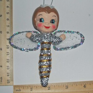 May include: A bee-themed ornament with a doll-like face, brown hair, and blue eyes. The body is covered in silver glitter with gold stripes and white glittery wings. A ruler shows the ornament is approximately 11.5 cm tall and 11.5 cm wide.